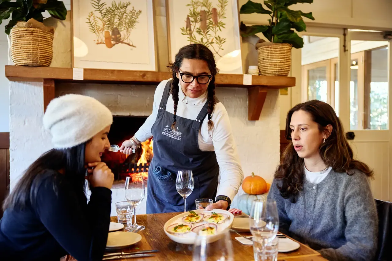 A staff member serves a plate of scallops to two diners at a table by the open fire at Merricks General Wine Store