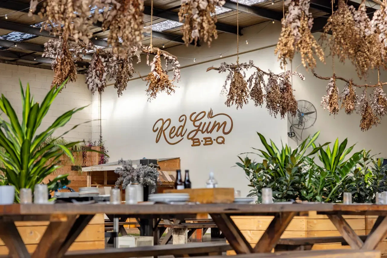 Dried flowers hang from the ceiling above long timber tables and potted plants, with the Red Gum BBQ sign behind
