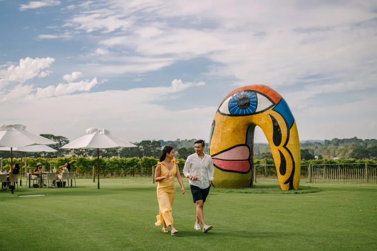 Two visitors stroll across the lawn at Pt. Leo Estate, with umbrella-shaded tables and a large mosaic face sculpture behind