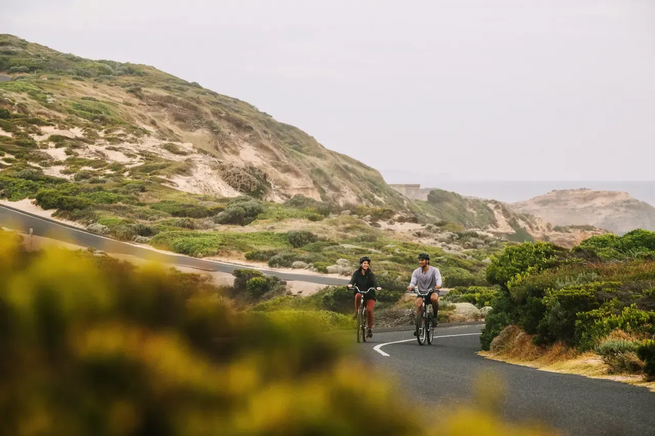 Two cyclists ride towards the camera on a quiet road through the dunes and coastal scrub of Point Nepean on an overcast day