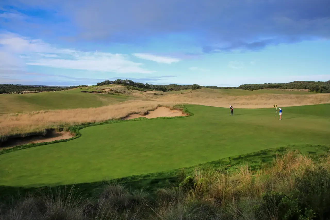Green fringed by sand bunkers and tussock grass at St Andrews Beach Golf Course, with two distant golfers near the flag