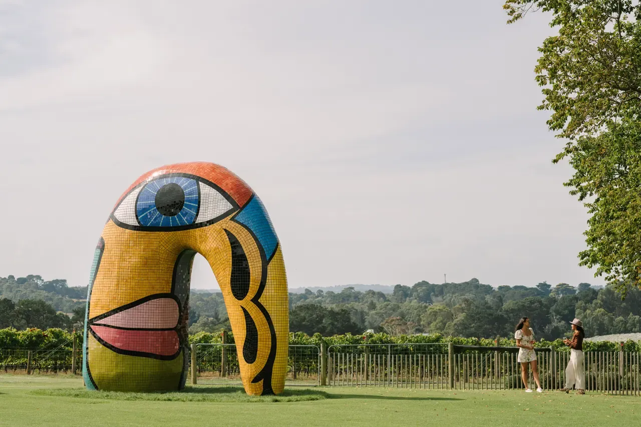 A colourful mosaic face sculpture shaped like an arch stands on the lawn at Pt. Leo Estate, with vineyards and bushland beyond