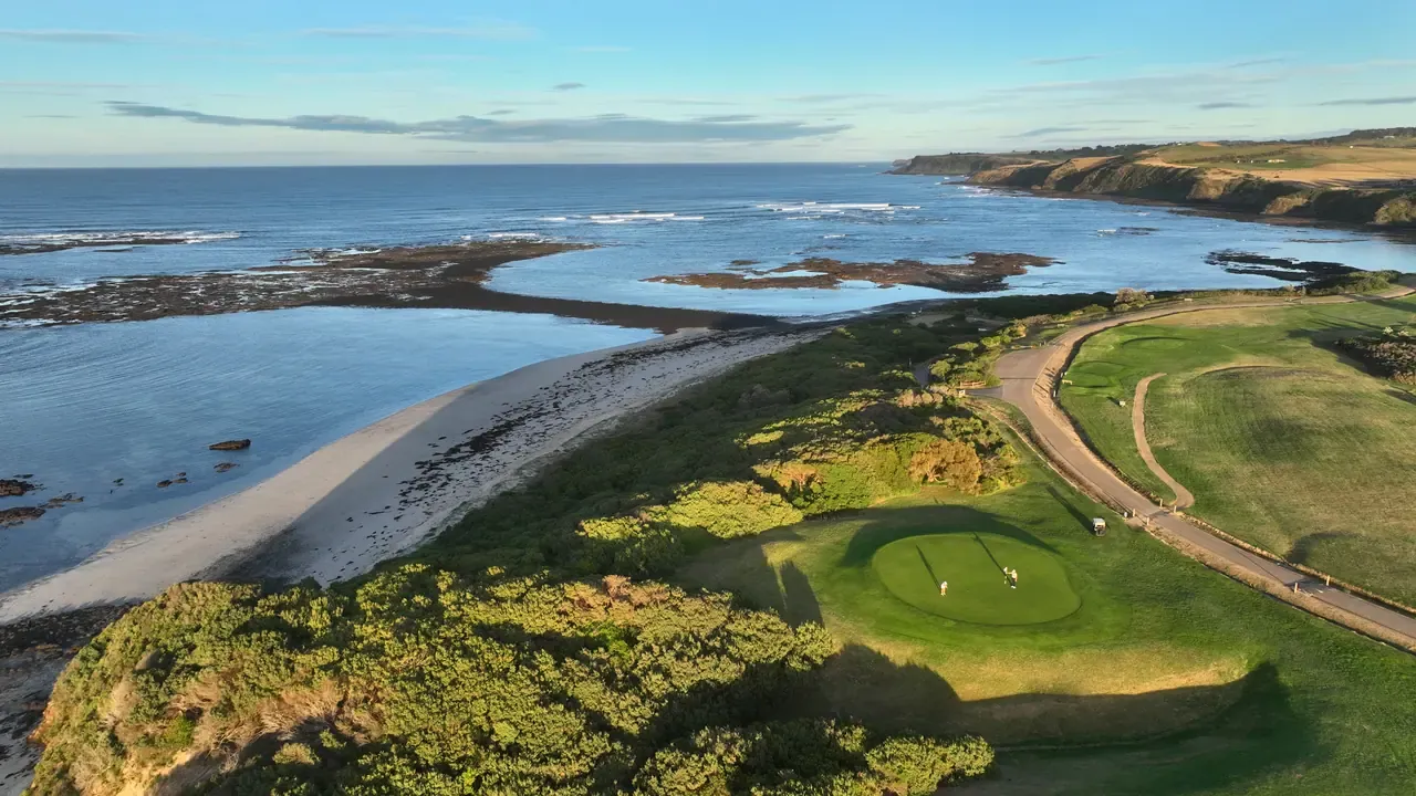 Aerial view of a coastal golf green on a headland, with two golfers, a sandy cove and rocky reef platforms in warm evening light.
