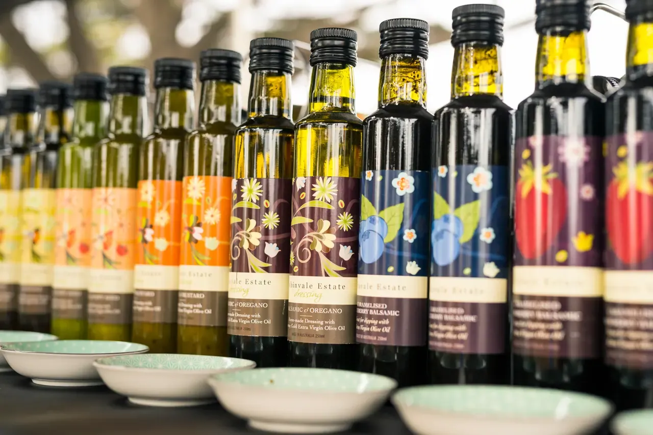 A row of bottled olive oils and dressings with floral labels behind small tasting dishes at a market stall