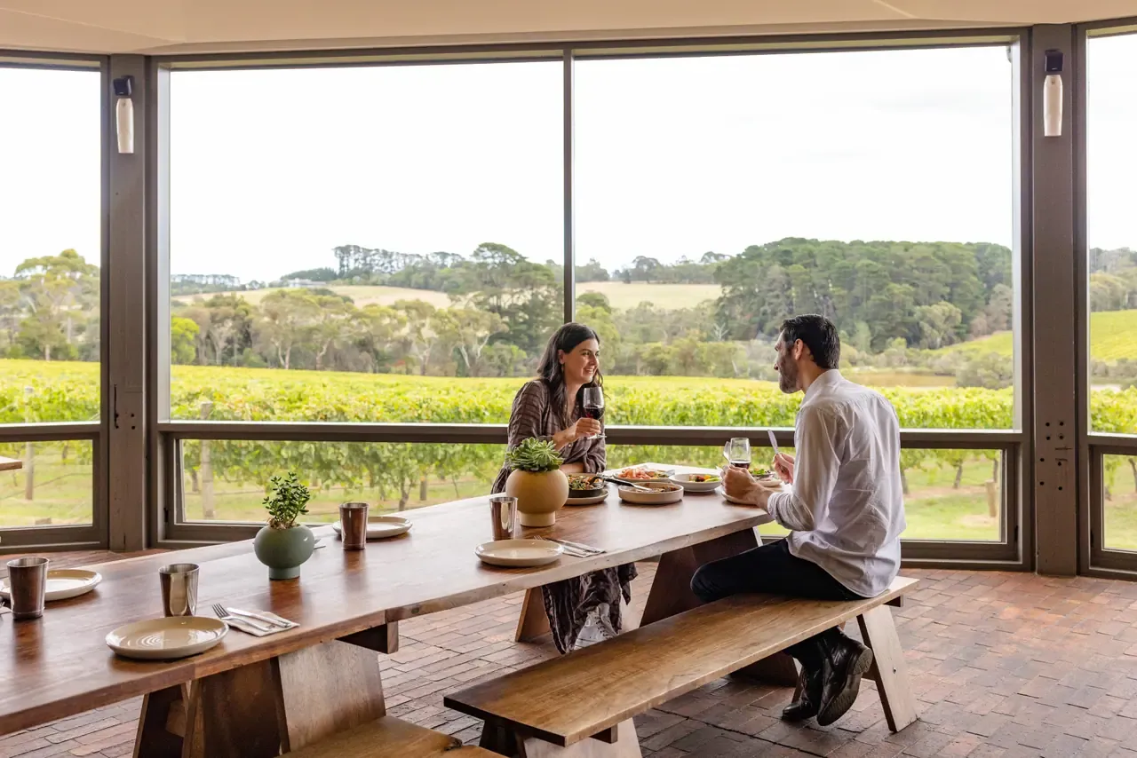 Two diners share plates and red wine at a long timber table beside floor-to-ceiling windows overlooking vineyard and bushland