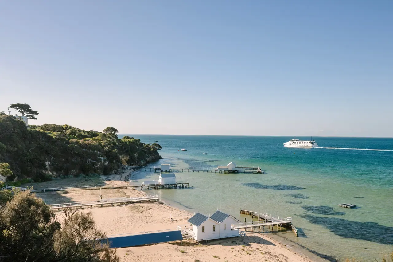 White boat sheds and jetties on a sandy beach below Millionaire's Walk, with a ferry crossing clear turquoise water