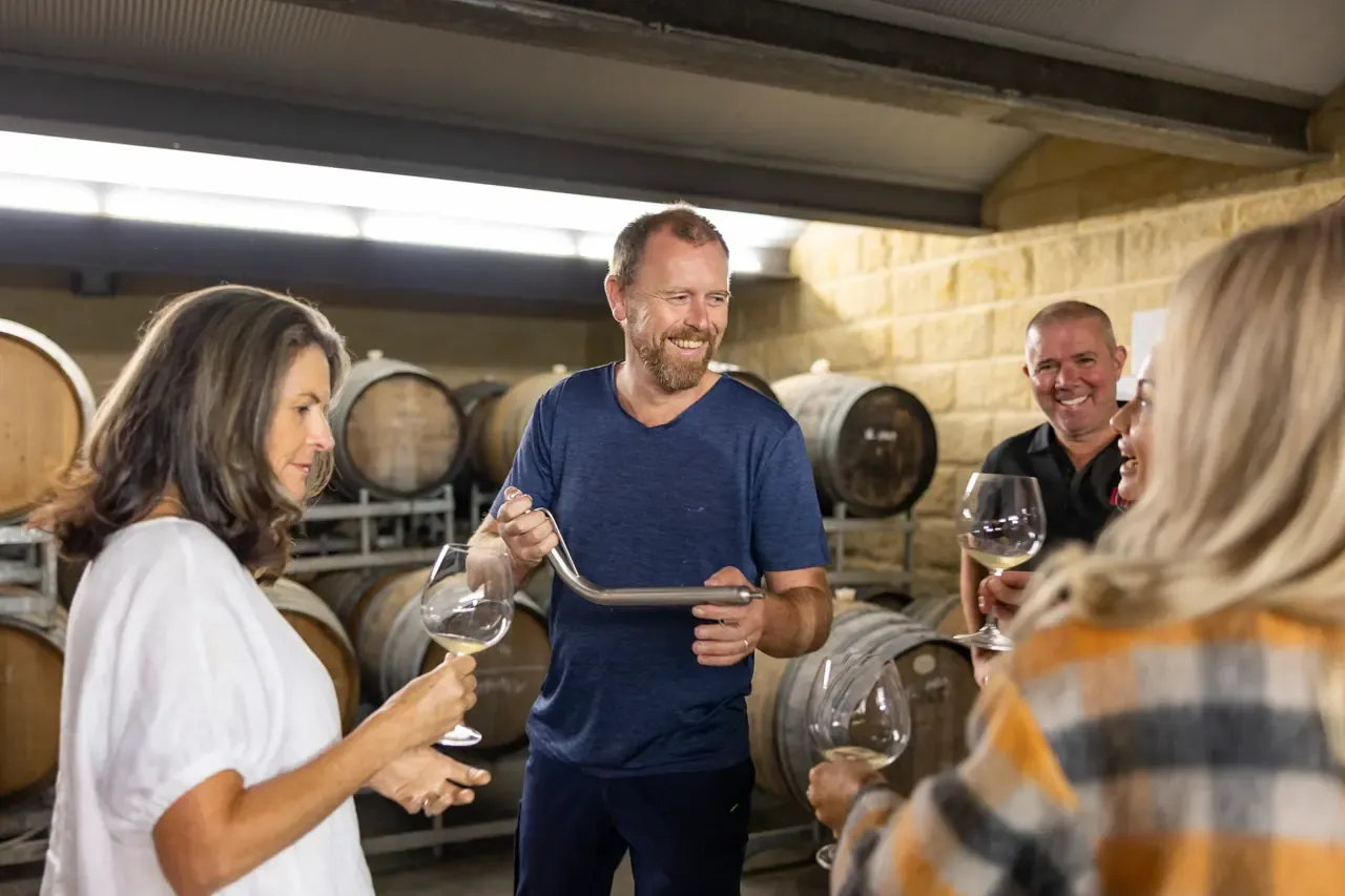 A winemaker draws wine from a barrel with a wine thief for smiling guests in the Ocean Eight barrel room