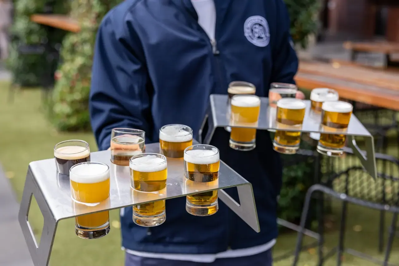 A staff member carries two steel tasting paddles holding glasses of dark, amber and pale beer across a garden