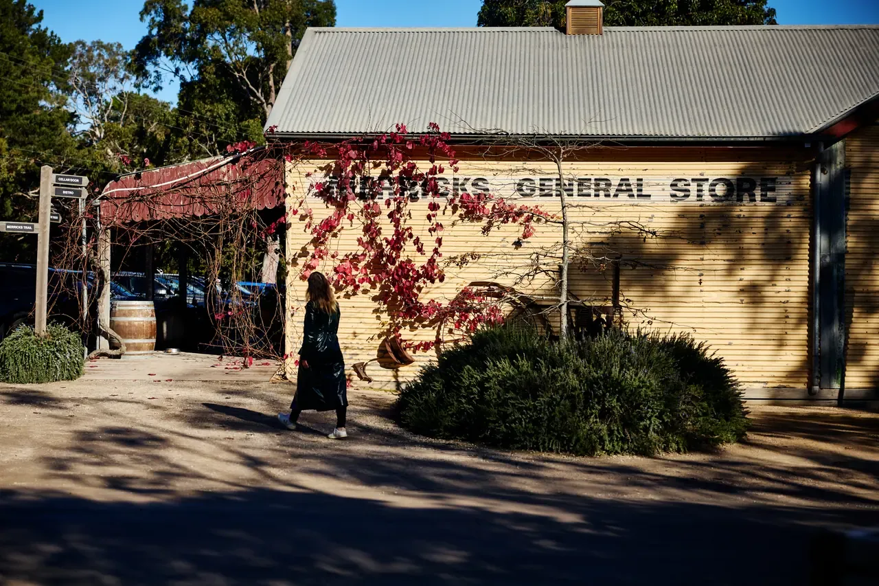 A visitor walks past the weatherboard facade of Merricks General Store, its painted sign framed by a red flowering vine