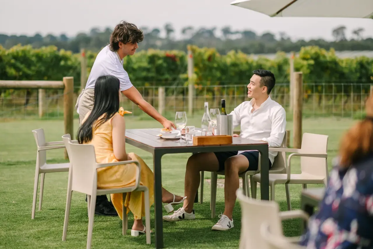 A server attends to two guests at an outdoor table on the lawn at Pt. Leo Estate, with vineyard rows behind