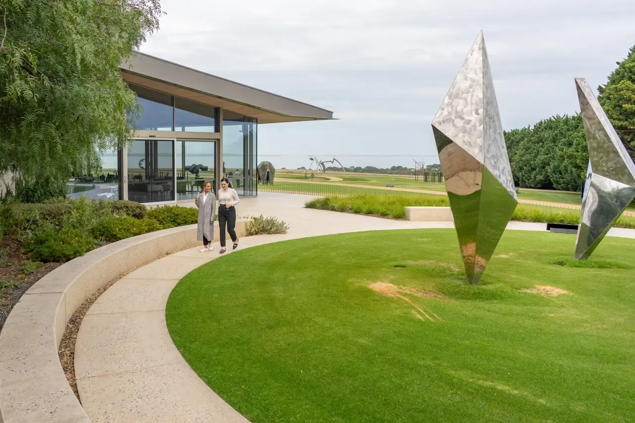 Two people walking a curved path past mirrored steel sculptures outside the glass-fronted Pt. Leo Estate building