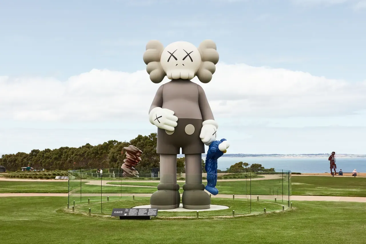 Giant grey KAWS figure holding a small blue figure on the sculpture park lawn at Pt. Leo Estate, with the sea and other artworks behind