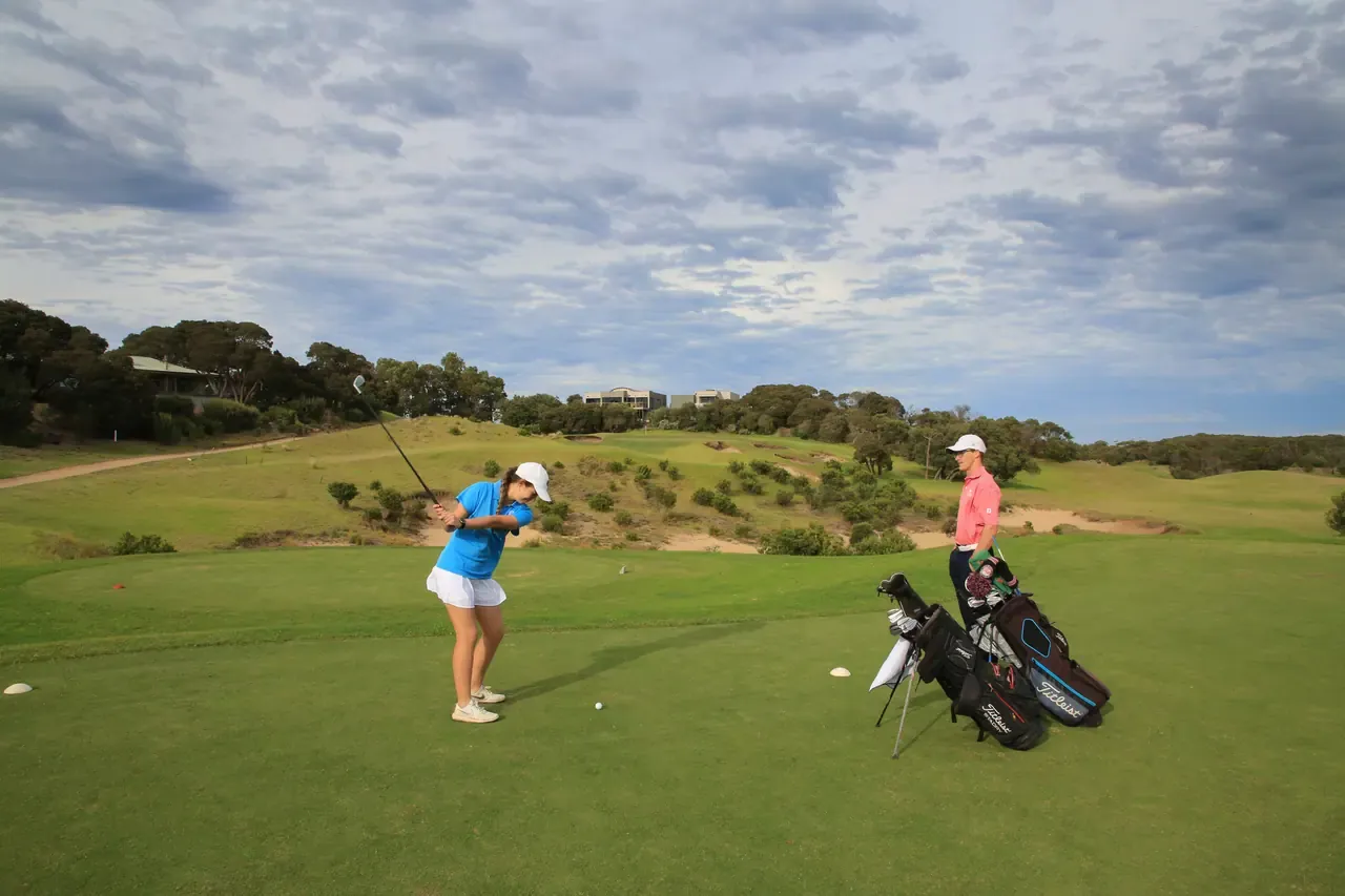 A golfer plays a shot from the tee at Moonah Links while a partner watches beside the bags, rolling scrubby dunes behind