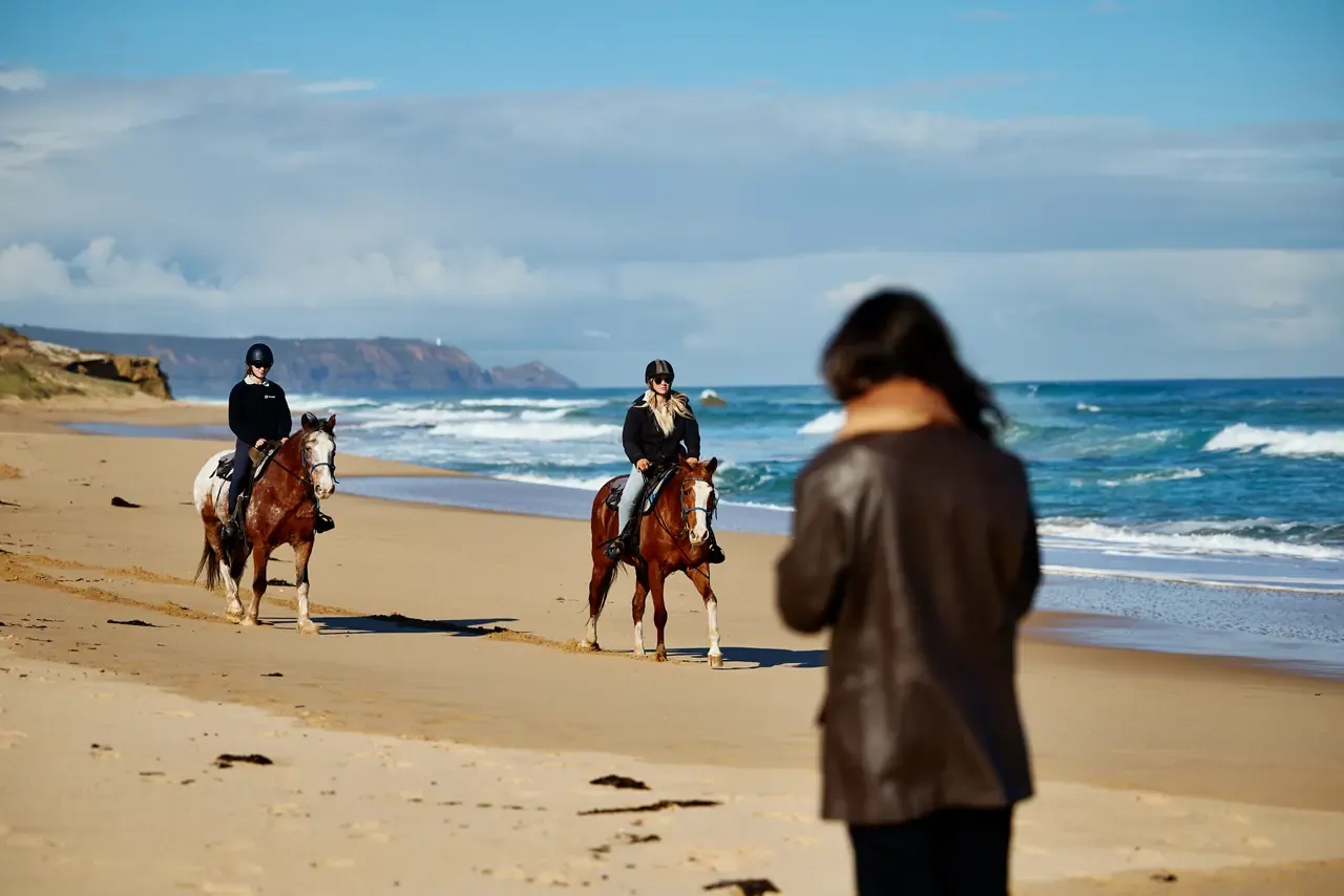 Two riders on horseback approach along Gunnamatta Beach as a blurred figure in a leather jacket stands in the foreground
