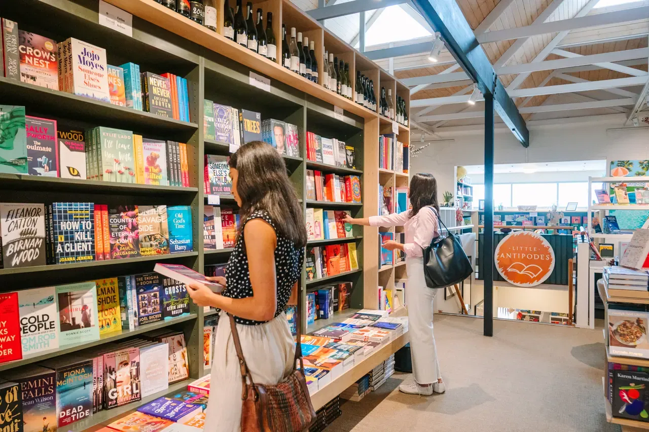 Two women browse tall shelves of books and wine under the timber-lined ceiling of Antipodes Bookshop in Sorrento
