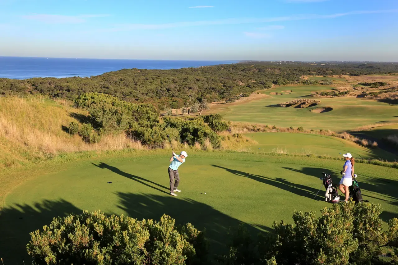 A golfer tees off at The National Golf Club with fairways and bunkers below, and the ocean beyond a band of coastal scrub