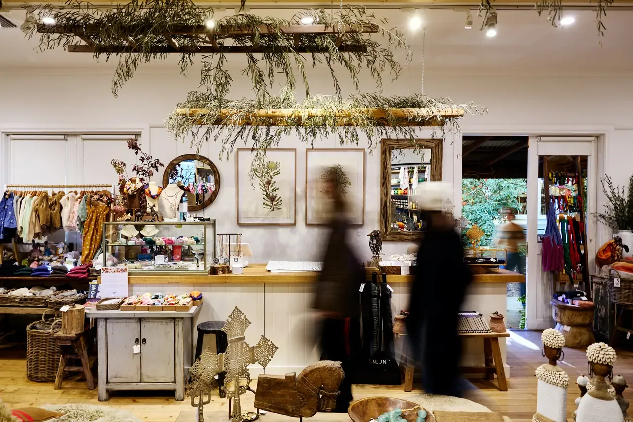 Shoppers pass through a homewares store with clothing racks, botanical prints and hanging dried foliage at Merricks General Wine Store