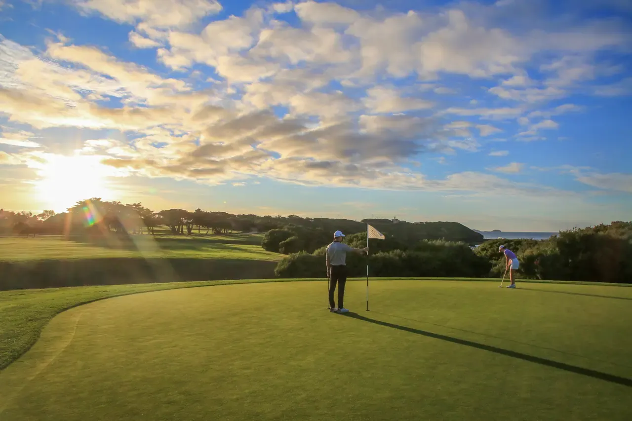 A golfer holds the flag on a green at Flinders Golf Course as another lines up a putt, low sun breaking through cloud