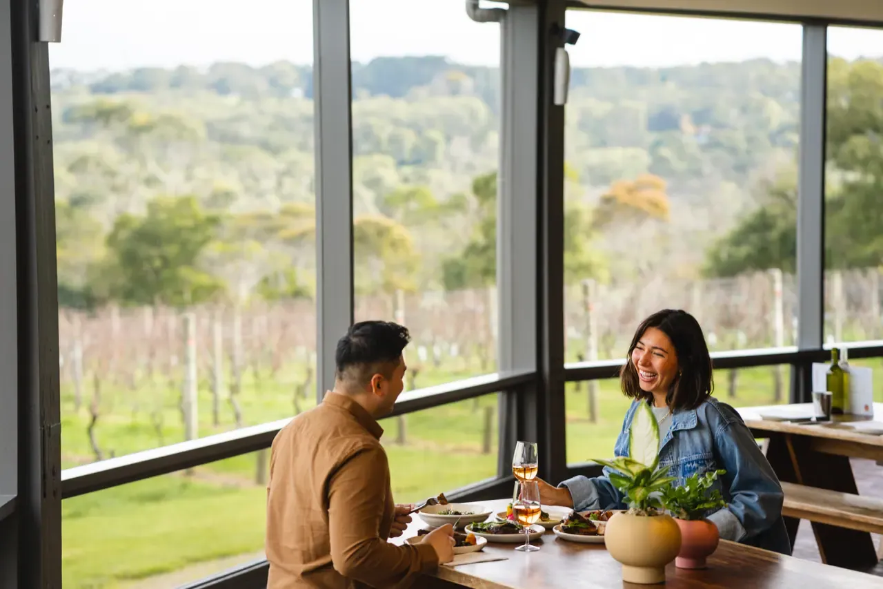 Two diners share plates and wine at a window table at Rare Hare, looking out over vineyard rows and bushland