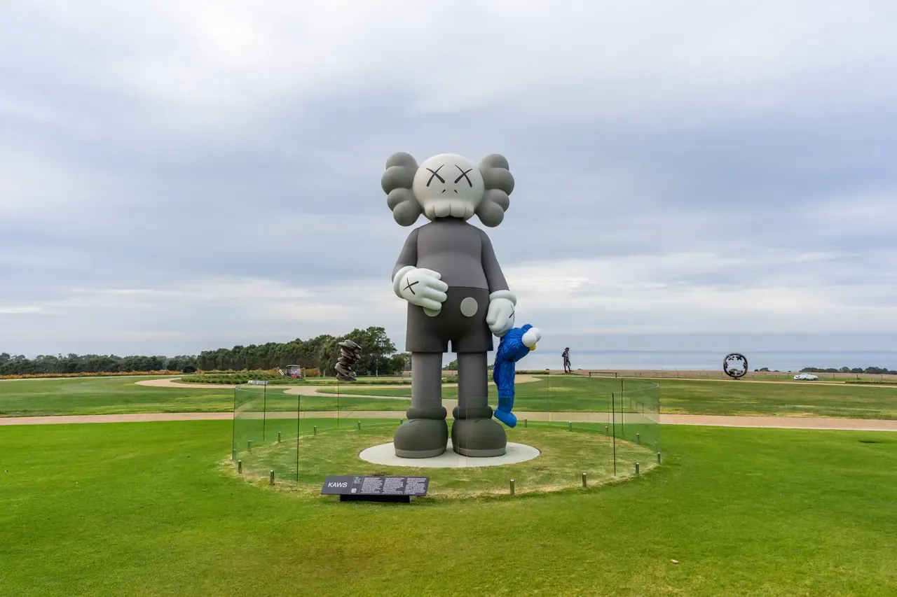 Giant grey KAWS figure holding a small blue figure on a lawn at Pt. Leo Estate sculpture park, with the sea beyond