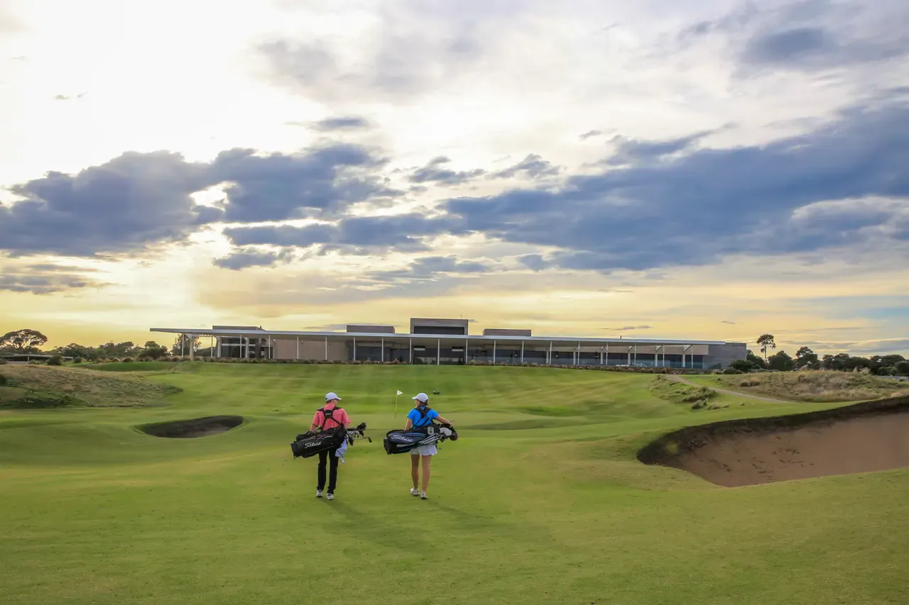 Two golfers carrying bags walk a fairway between bunkers towards the clubhouse at Moonah Links Golf Course under a dramatic evening sky.