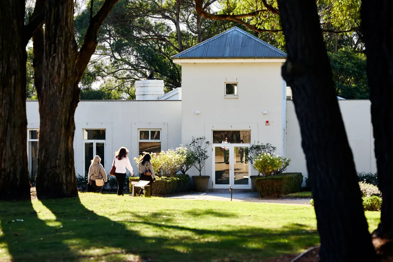 Visitors cross a sunlit lawn between large tree trunks towards the white homestead building at Lindenderry at Red Hill