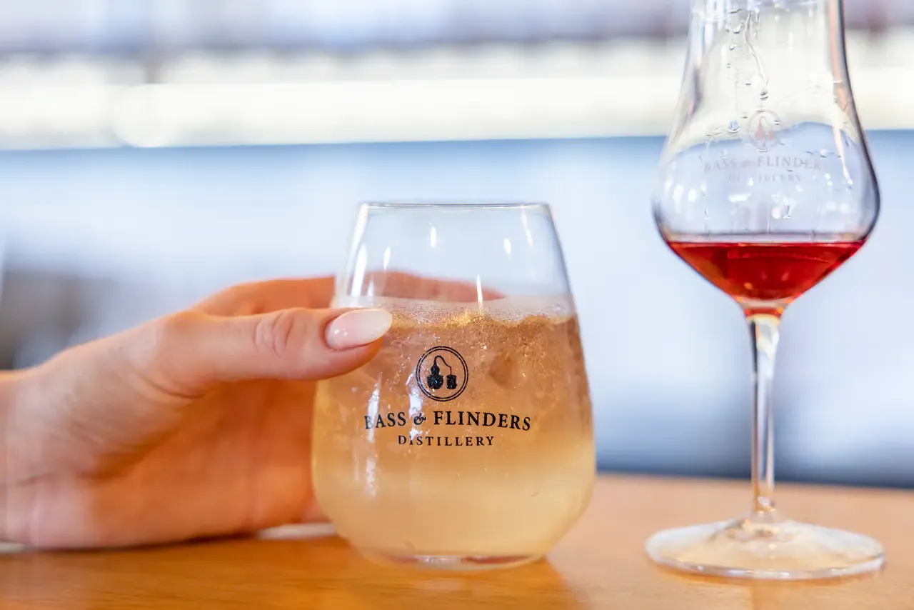A hand holds a Bass and Flinders Distillery tumbler of gin and tonic on the bar beside a stemmed glass of pink spirit