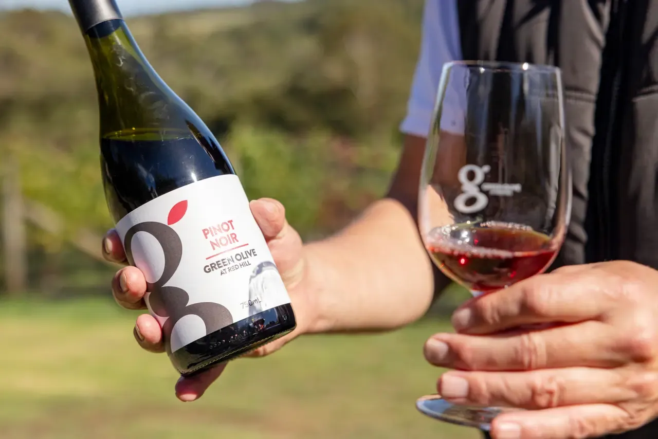 A hand holds a bottle of Green Olive at Red Hill Pinot Noir beside a glass of red wine, with farmland blurred behind