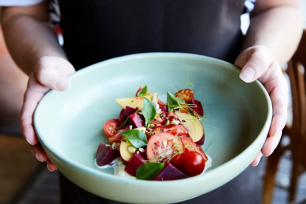 Hands holding a pale green bowl of beetroot, cherry tomato and fruit salad topped with herbs and sprouts