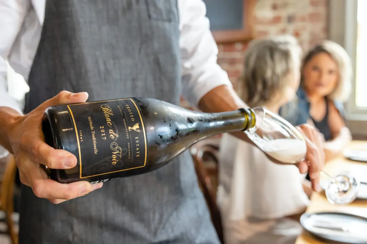Staff member pouring a Trofeo Estate 2017 Blanc de Noir sparkling wine into a glass, with diners blurred behind