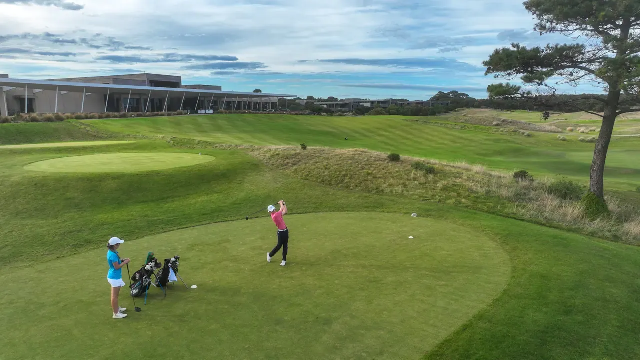 A golfer drives from the tee while a partner waits beside two golf bags, with a low modern clubhouse and pine tree in the background.