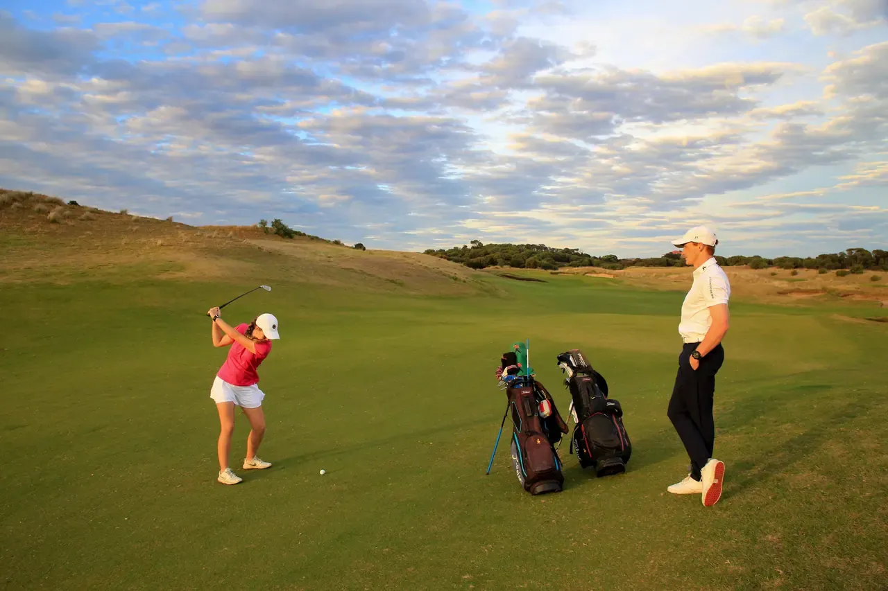 A golfer takes a swing on the fairway at The Dunes Golf Links while a second player stands beside two golf bags in warm light