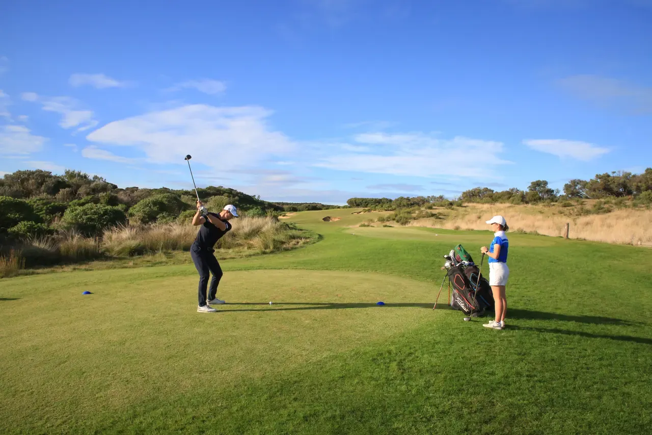 A golfer swings from an elevated tee at St Andrews Beach Golf Course while a partner waits by the bags, the fairway rolling ahead