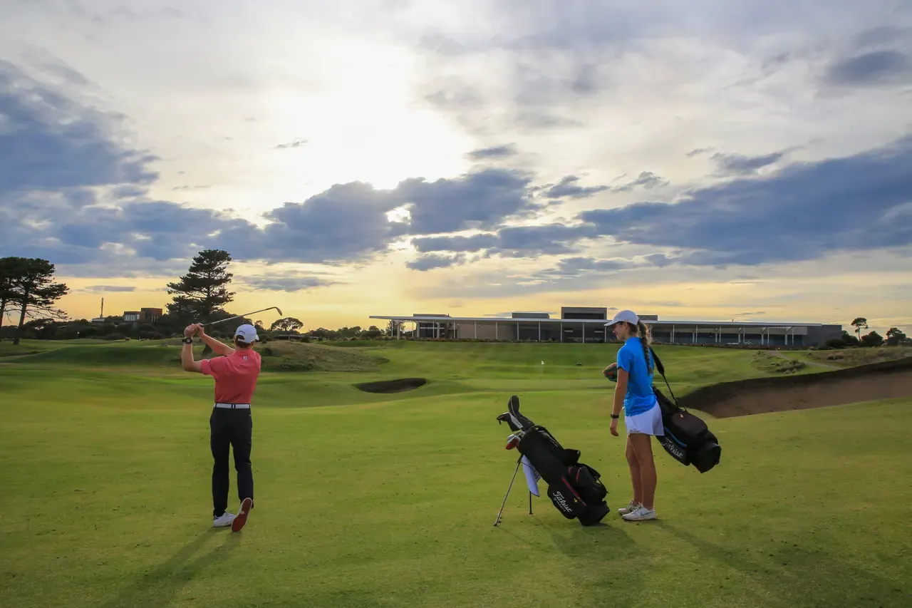 A golfer follows through on a fairway at Moonah Links as a partner waits by a bag, the clubhouse under a glowing evening sky
