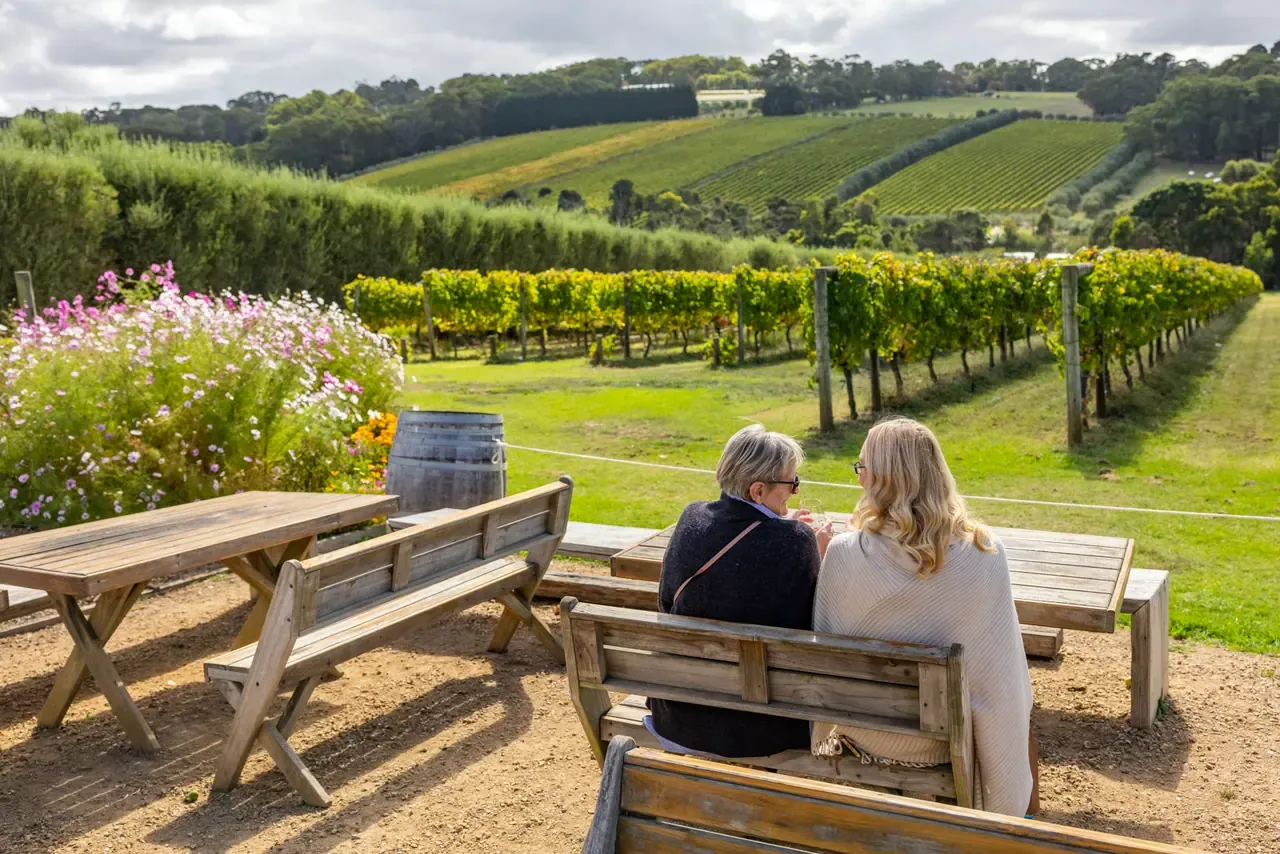 Two people seated on a timber bench looking over rows of vines and hillside vineyards at Montalto, with flowers to one side