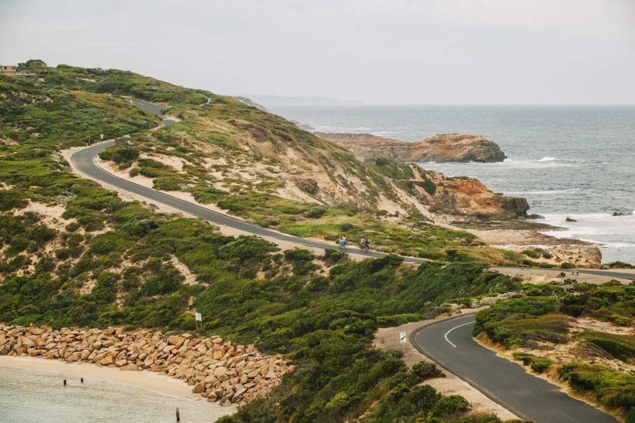 Two cyclists on a road winding through coastal scrub and sandy dunes at Point Nepean above rocky headlands and a grey sea