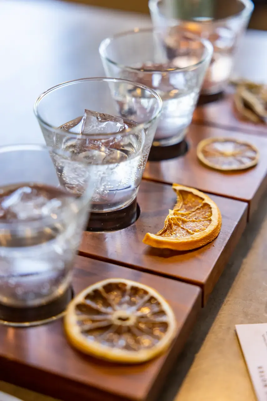A tasting paddle of gin and tonics over ice on a timber board, each glass paired with a slice of dried orange