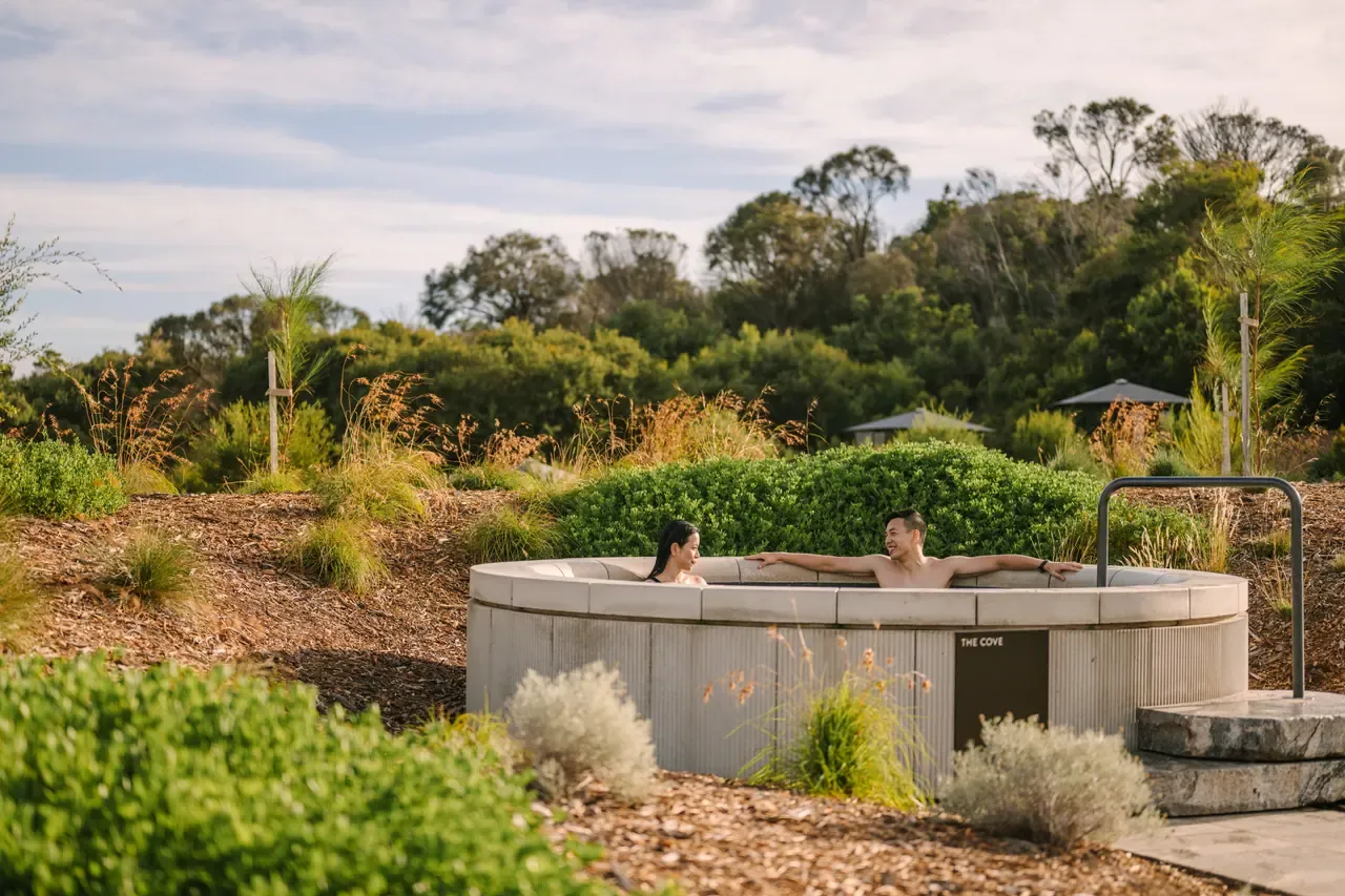 Two bathers relaxing in a round concrete hot pool set among native coastal gardens at Alba Thermal Springs