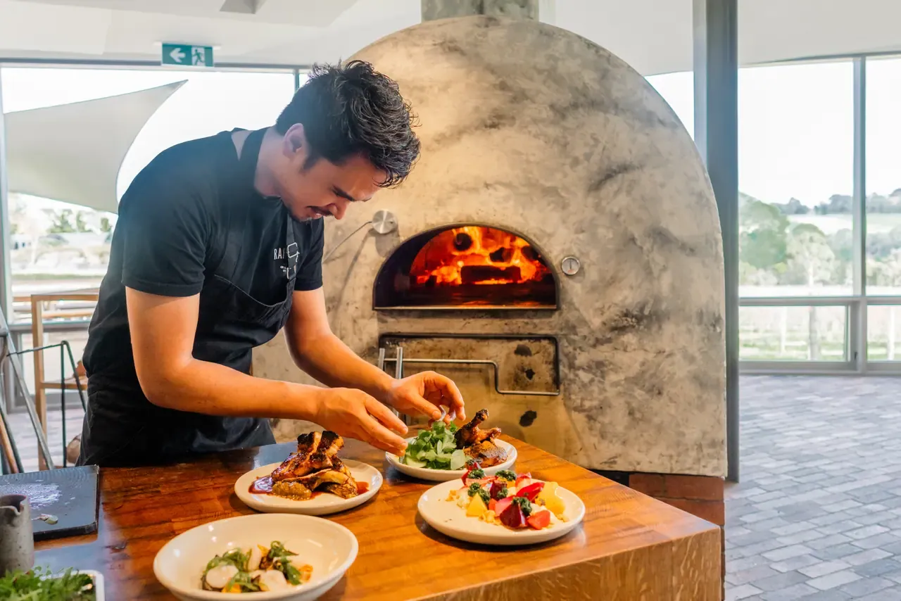 A chef garnishes plates of roast meat and salad in front of a glowing wood-fired oven in a light-filled kitchen