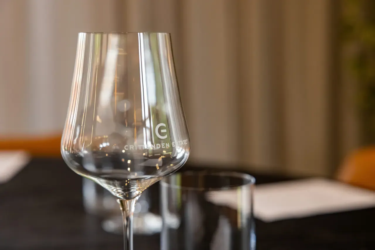 An empty wine glass etched with the Crittenden Estate logo stands on a dark table beside a water tumbler