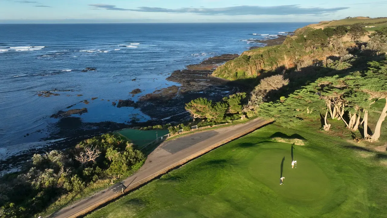 Aerial view of two golfers on a seaside green beside a cart path, with cypress trees and rock shelves along the Flinders coastline.