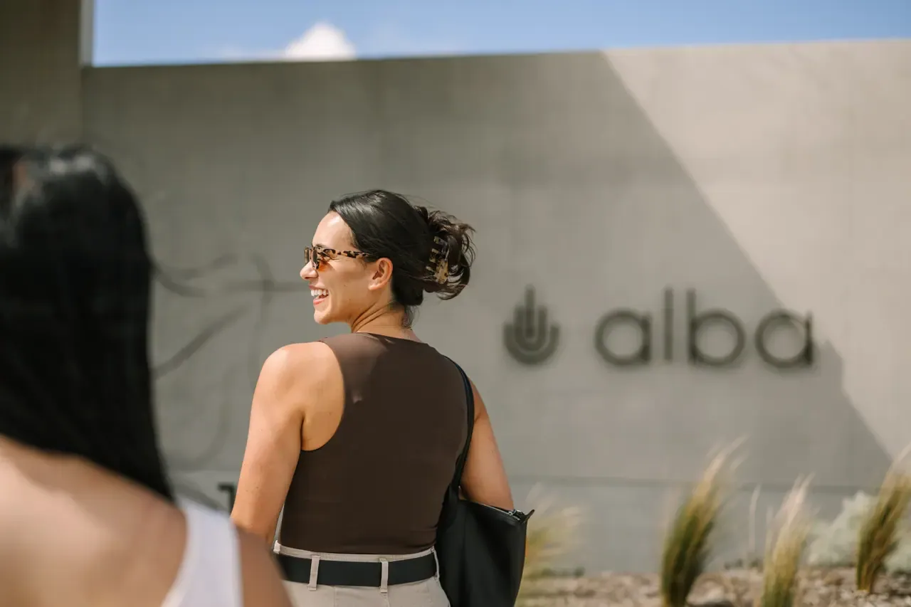 Smiling visitor in sunglasses walks past a concrete wall bearing the Alba logo and name, with ornamental grasses in the foreground