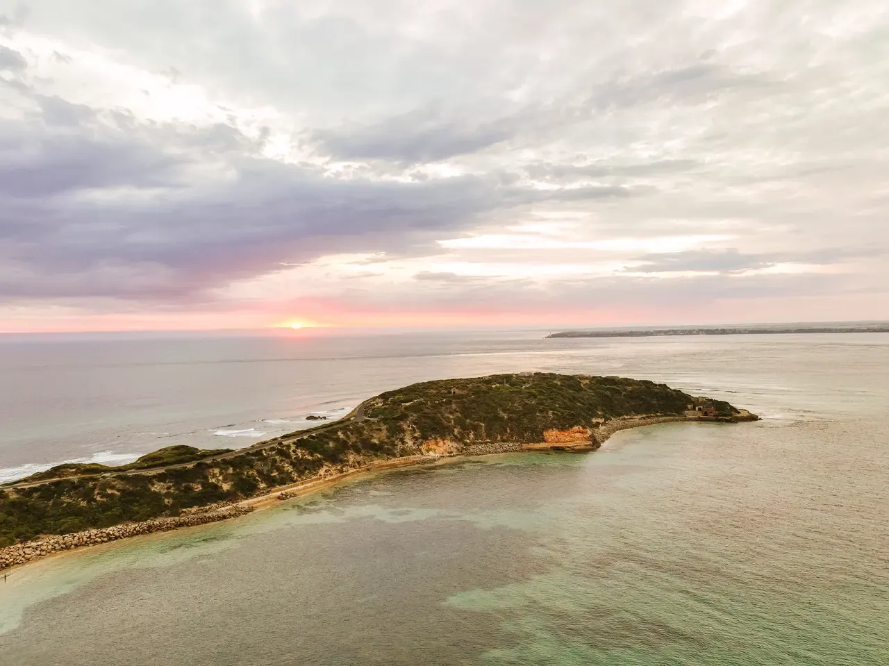 Aerial view of the narrow scrubby tip of Point Nepean reaching into calm water, with a pink and lilac sunset sky over the horizon.