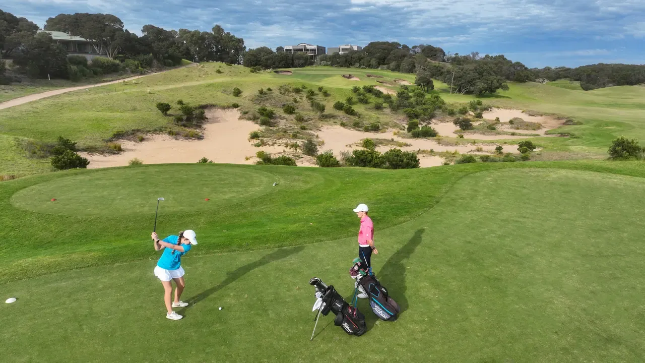 Aerial view of a golfer mid-swing from the tee at Moonah Links, with a second golfer, bags on the grass and sandy dunes behind.