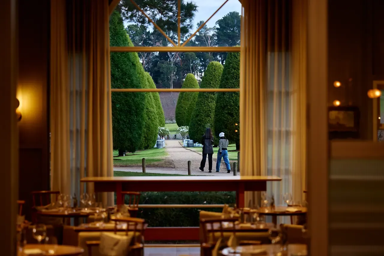 View from a set dining room through a tall window to two people walking a gravel path between clipped conical cypress trees