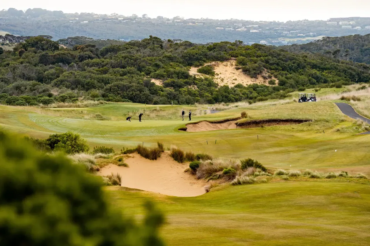 Golfers on a distant green framed by sand bunkers and dune scrub at St Andrews Beach Golf Course, with a buggy on the cart path