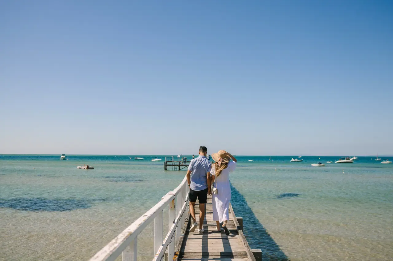 A couple walks along a narrow white-railed pier at Sorrento over clear shallow water dotted with moored boats