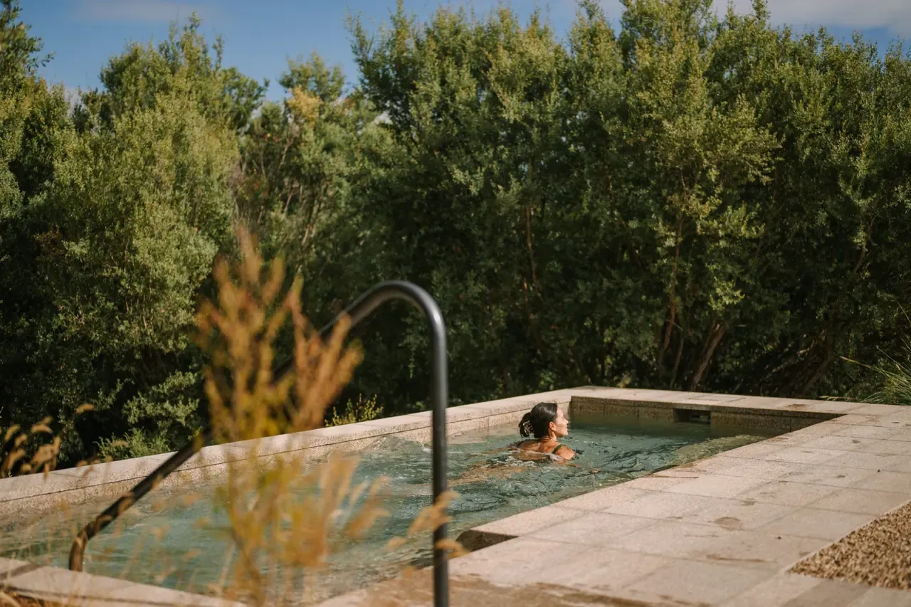 A bather swimming across a stone-edged plunge pool backed by dense coastal trees at Alba Thermal Springs
