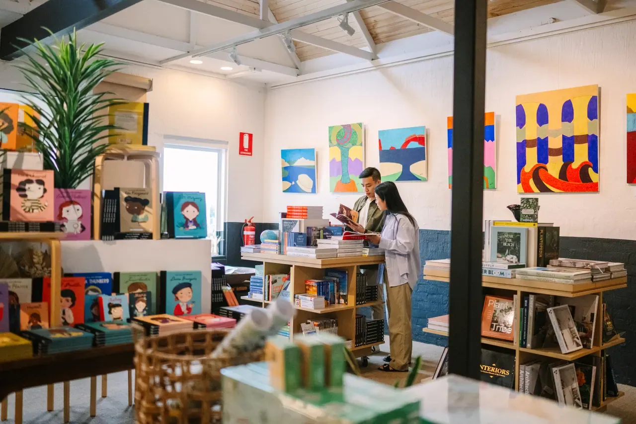 A couple reads together among low book tables and colourful paintings in the gallery space at Antipodes Bookshop, Sorrento