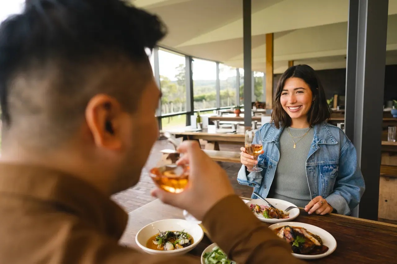 A diner smiles with a glass of rose over shared plates in a dining room with windows onto vineyard hills